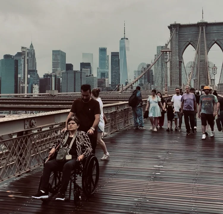 Persona en silla de ruedas siendo empujada por un acompañante sobre el Puente de Brooklyn con el skyline de Nueva York al fondo