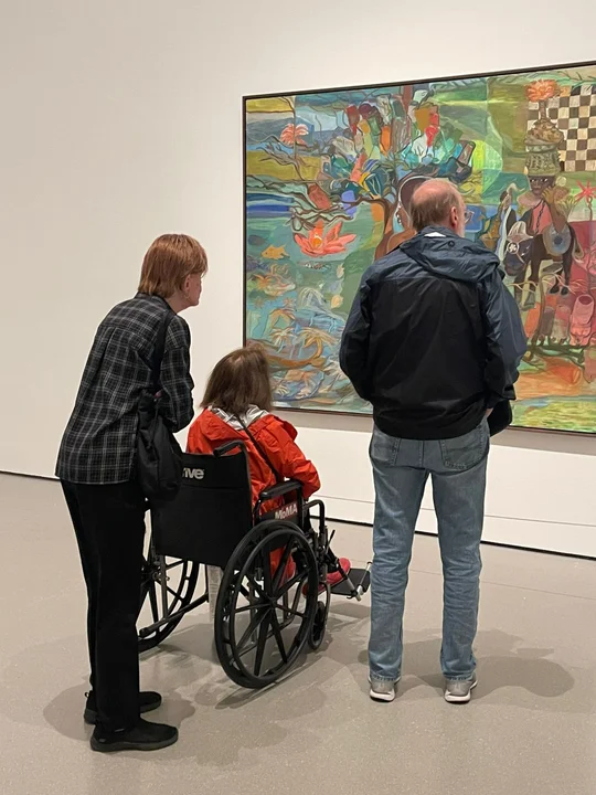 Persona en silla de ruedas observando una pintura en un museo junto a dos acompañantes