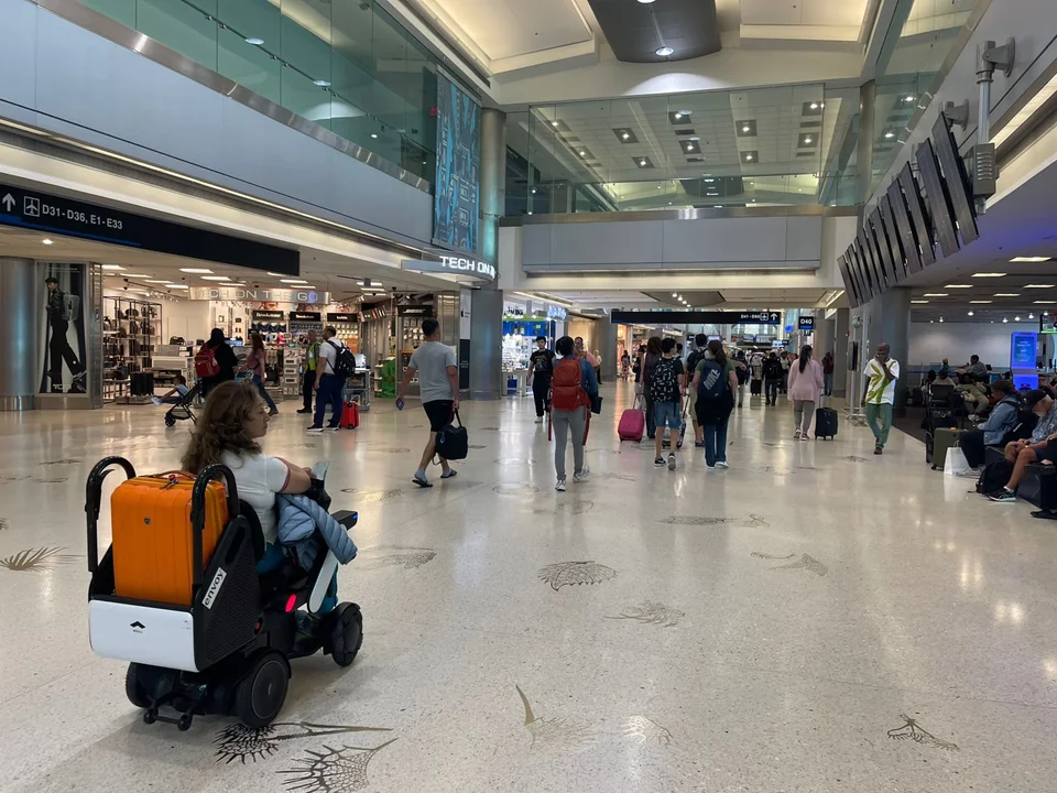 Mujer en silla de ruedas eléctrica con una valija naranja y otra blanca desplazándose por un aeropuerto moderno, rodeada de viajeros con equipaje de mano y señalización visible hacia las puertas de embarque.