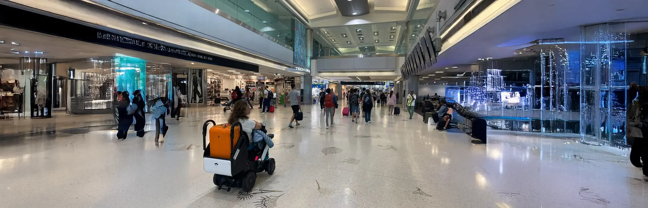 Mujer en silla de ruedas eléctrica con una valija naranja y otra blanca desplazándose por un aeropuerto moderno, rodeada de viajeros con equipaje de mano y señalización visible hacia las puertas de embarque.