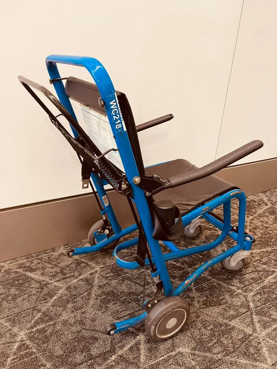 Silla de ruedas azul de transporte utilizada en aeropuertos para asistir a pasajeros con movilidad reducida, apoyada sobre una alfombra gris junto a una pared blanca.