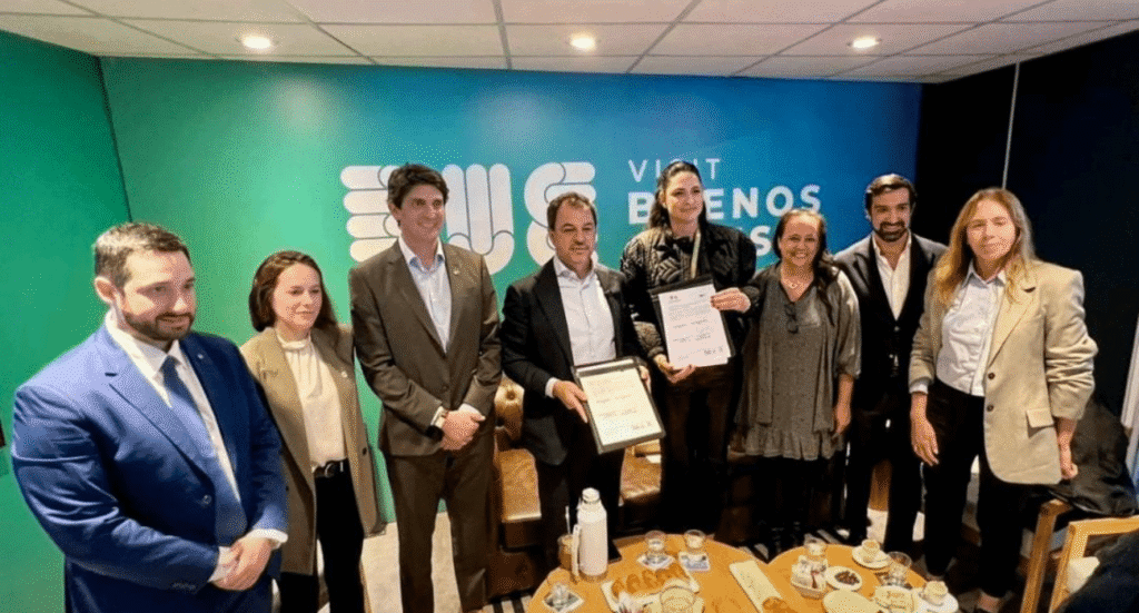 Grupo de ocho personas posando en una sala de reuniones, algunas sosteniendo certificados, frente a un fondo azul con el texto “Visit Buenos Aires”. En la mesa hay tazas, termos y platos con facturas.